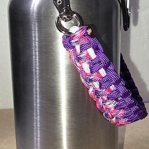 May include: A stainless steel water bottle with a black lid and a braided purple, pink, and white strap. The bottle has a silver carabiner clip attached to the strap.