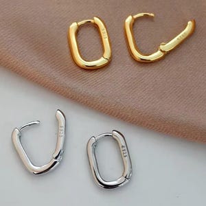 May include: Two pairs of hoop earrings. One pair is gold-coloured, the other is silver-coloured. Each earring has a rectangular shape with a hinged closure. The earrings are displayed on a neutral-toned fabric.