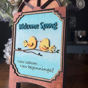Puede incluir: Cartel de madera con fondo azul claro y las palabras "Welcome Spring" en turquesa. Dos pájaros amarillos se posan en una rama. El cartel también dice "New season... New beginnings!" en negro. El marco es de color marrón oscuro.