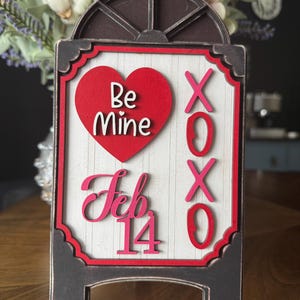 以下が含まれることがあります： 「Be Mine」と「Feb 14」と書かれた赤いハートの木製バレンタインデーのサイン。サインにはピンク色の「XOXO」も表示されています。サインは黒いフレームと白い背景です。