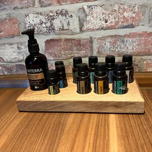 Puede incluir: Un expositor de madera para aceites esenciales que contiene varias botellas pequeñas de vidrio marrón con tapas negras. Una botella marrón más grande con un dispensador de bomba está a la izquierda. Las botellas están etiquetadas con varios nombres de aceites esenciales.
