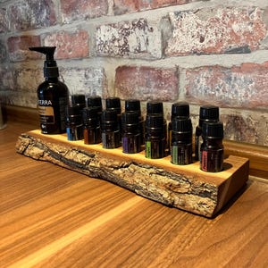 Könnte beinhalten: Ein Holzregal für ätherische Öle mit einer rohen Kante, das mehrere kleine dunkle Flaschen enthält. Auf der linken Seite befindet sich eine größere Flasche mit einem Pumpspender. Das Regal steht auf einer Holzoberfläche, mit einer Backsteinmauer im Hintergrund.