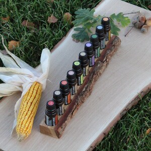 May include: A wooden log with slots to hold 12 small bottles of essential oils. The bottles are various colors and have labels with the word "doTERRA" on them. The log is sitting on a wooden board with a corn cob and husk on the left side.