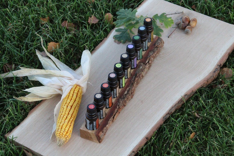 May include: A wooden log with slots to hold 12 small bottles of essential oils. The bottles are various colors and have labels with the word "doTERRA" on them. The log is sitting on a wooden board with a corn cob and husk on the left side.