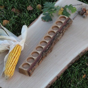May include: A wooden tray with 12 round indentations, made from a tree branch, sits on a wooden slab. An ear of corn and acorns are also on the slab.