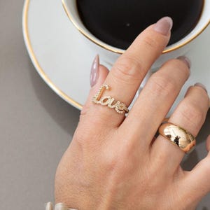 May include: A hand wearing two gold rings, one spelling "love" with small stones, the other a simple band. A white teacup with a gold rim and dark coffee sits nearby. The sleeve of a beige blouse is visible.