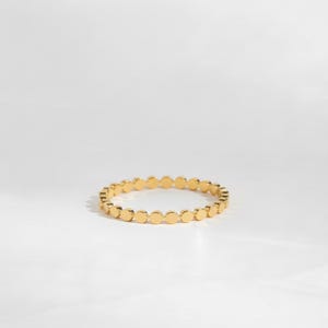 May include: A gold-colored ring with a series of small, flat, circular accents. The ring is set against a white background, with soft shadows adding depth. The ring's design is simple and elegant, suitable for everyday wear.