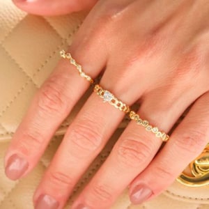 Puede incluir: Una mano que luce tres anillos de oro con detalles de diamantes. Los anillos presentan diferentes estilos, incluyendo un diseño de cadena delicada, un anillo con un diamante central y un anillo con pequeños diamantes espaciados. El fondo es un bolso beige acolchado.