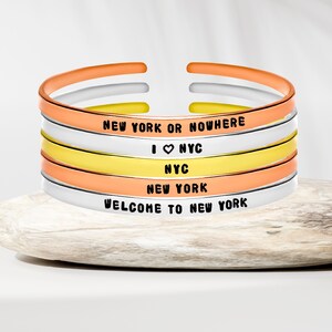 Può includere: Pila di braccialetti a polsino aperti nei toni dell'arancione, bianco e giallo. Ogni braccialetto è inciso con una frase diversa su New York City, tra cui "NEW YORK OR NOWHERE" e "WELCOME TO NEW YORK."