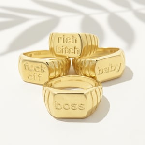 Op de afbeelding: Vier goudkleurige zegelringen met reliëftekst. De ringen tonen de woorden "rich bitch", "fuck off", "baby" en "boss". De ringen hebben een rechthoekige voorkant met verticale ribbels aan de zijkanten.