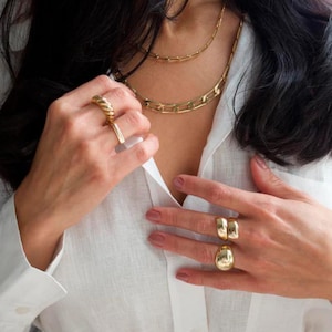 May include: Gold-toned jewelry set featuring a chain necklace and multiple rings. The rings include a twisted design, a simple band, and two chunky styles. The jewelry is worn with a white button-down shirt.