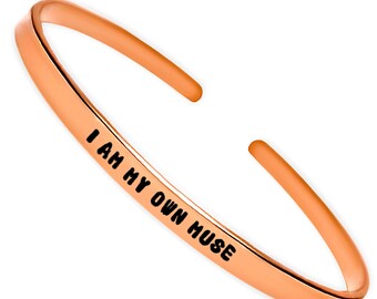 May include: A rose gold bangle bracelet with the words "I AM MY OWN MUSE" engraved on it.