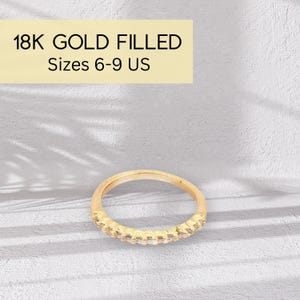 May include: An 18K gold-filled ring with a band of small, clear stones. The ring is shown against a light background. Text in the image reads "18K GOLD FILLED Sizes 6-9 US".