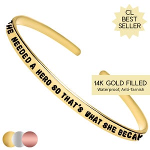 May include: Gold-filled bangle bracelet with the inscription "HE NEEDED A HERO SO THAT'S WHAT SHE BECAME".