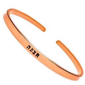 May include: A rose gold bangle bracelet with the Hebrew word "Mazel Tov" engraved on it.