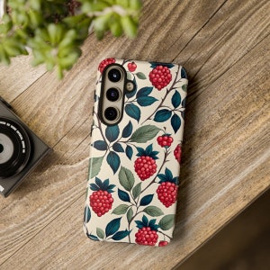 K&ouml;nnte beinhalten: Eine Handyh&uuml;lle mit cremefarbenem Hintergrund und einem Muster aus roten Himbeeren, gr&uuml;nen Bl&auml;ttern und dunkelblauen Stielen. Die H&uuml;lle hat einen schwarzen Rand und einen Kameraausschnitt. Die H&uuml;lle liegt auf einer Holzoberfl&auml;che.