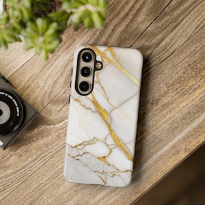 May include: A white phone case with a gold marble pattern. The case has a black camera cutout with three lenses.
