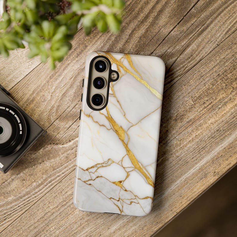 May include: A white phone case with a gold marble pattern. The case has a black camera cutout with three lenses.