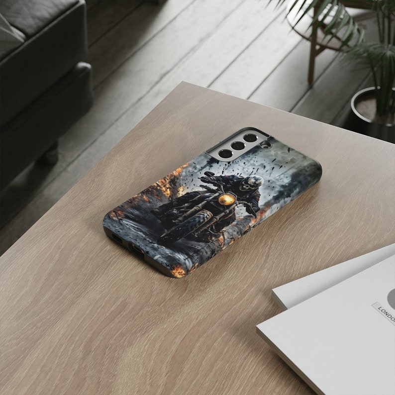 Puede incluir: Una funda de tel&eacute;fono negra con un gr&aacute;fico de una persona conduciendo una motocicleta a trav&eacute;s de un paisaje de fuego. La funda tiene un recorte para el objetivo de la c&aacute;mara.