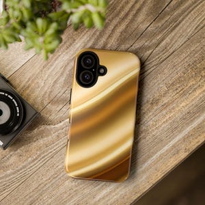 K&ouml;nnte beinhalten: Eine Smartphone-H&uuml;lle mit einem flie&szlig;enden, abstrakten Design in Gold- und Braunt&ouml;nen. Die H&uuml;lle hat einen Ausschnitt f&uuml;r eine Dual-Objektiv-Kamera und liegt auf einer Holzoberfl&auml;che. Eine Kamera und Gr&uuml;nzeug sind ebenfalls zu sehen.