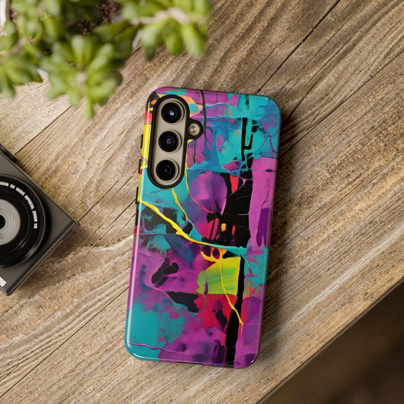 Op de afbeelding: Een telefoonhoesje met een abstract ontwerp in levendige kleuren zoals turquoise, paars, roze, geel en zwart. De hoes heeft een uitsparing voor een drievoudige camera lens.