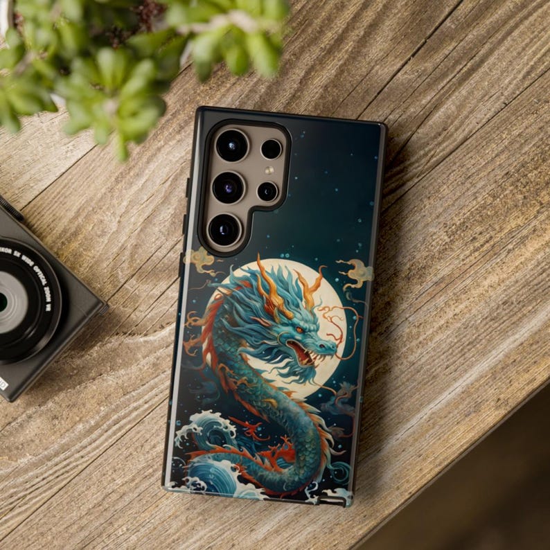 K&ouml;nnte beinhalten: Eine schwarze Handyh&uuml;lle mit einem blauen und gr&uuml;nen Drachen-Design. Der Drache ist von Wolken und einem Vollmond umgeben. Die Handyh&uuml;lle hat einen Ausschnitt f&uuml;r die Kamera.