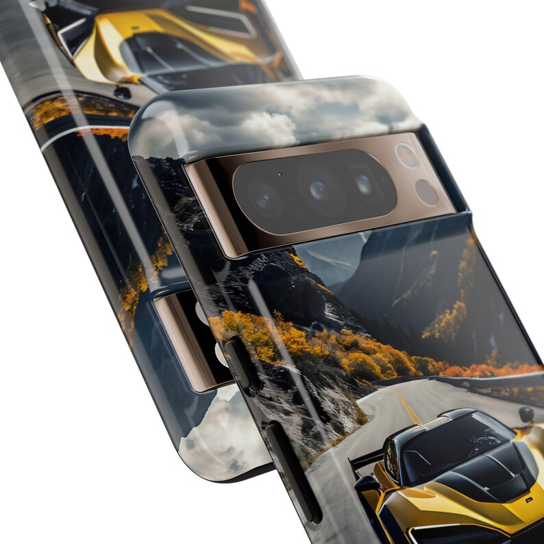 Puede incluir: Una funda para tel&eacute;fono con un coche deportivo amarillo conduciendo por una carretera de monta&ntilde;a sinuosa. La funda tiene una protuberancia de c&aacute;mara negra y dorada con tres lentes.