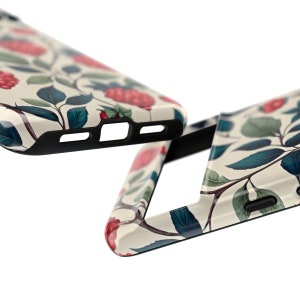 K&ouml;nnte beinhalten: Nahaufnahme von zwei Handyh&uuml;llen. Sie zeigen ein cremefarbenes Blumenmuster mit roten Beeren, gr&uuml;nen Bl&auml;ttern und dunkelbraunen Stielen. Die R&auml;nder der H&uuml;llen sind schwarz.