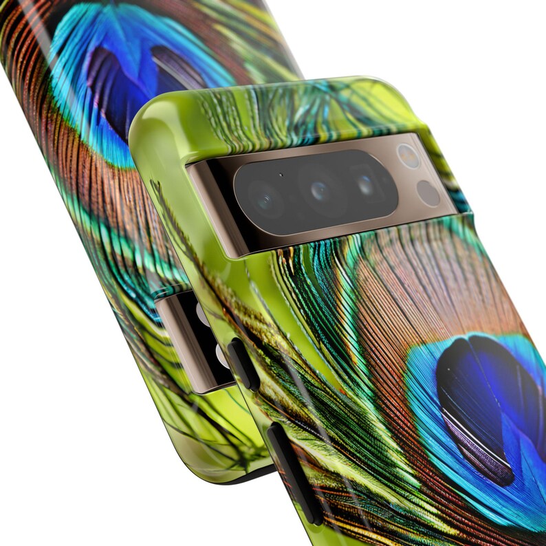 K&ouml;nnte beinhalten: Eine gr&uuml;ne, schwarze und blaue Handyh&uuml;lle mit einem Pfauenfeder-Design. Die H&uuml;lle hat eine erh&ouml;hte Kameraabdeckung mit drei Objektiven.