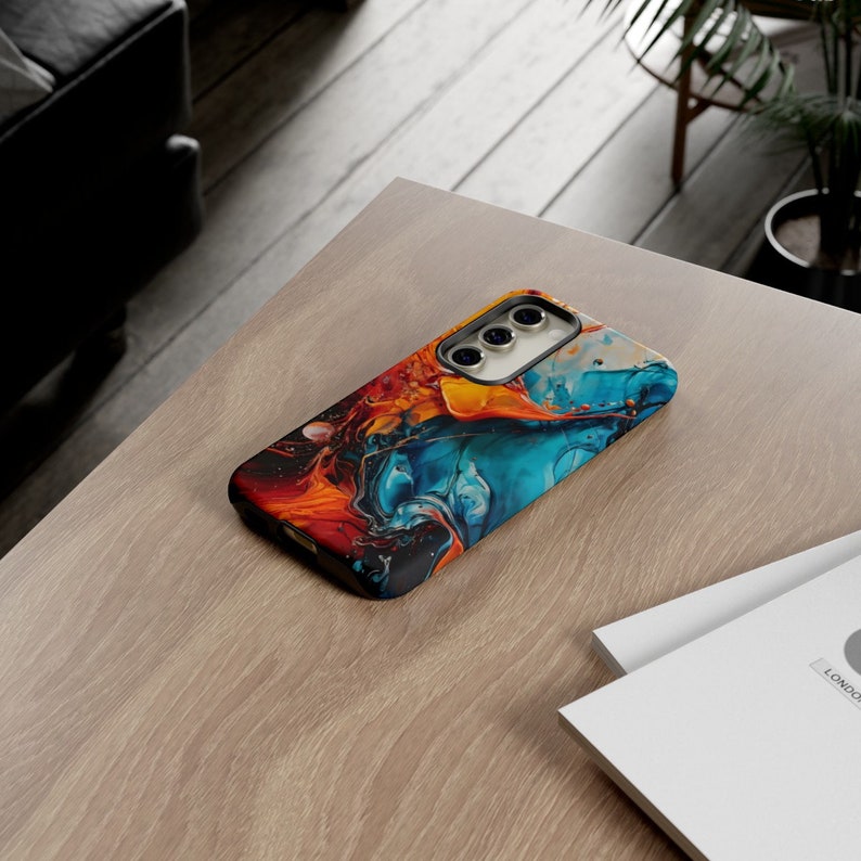 Puede incluir: Una funda para tel&eacute;fono con un dise&ntilde;o abstracto colorido que presenta remolinos de pintura azul, naranja y amarilla. La funda tiene un borde negro y un recorte para el objetivo de la c&aacute;mara.