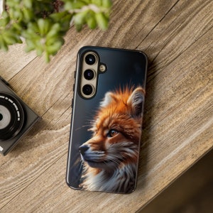 以下が含まれることがあります： 赤いキツネのイラストが描かれた黒い携帯電話ケース。ケースの背面にはトリプルカメラレンズがあります。