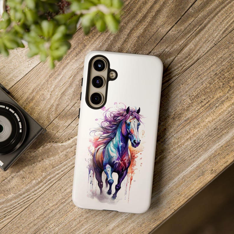 May include: A white phone case featuring a colourful watercolour painting of a horse running. The horse is purple, blue and orange with a white mane and tail.