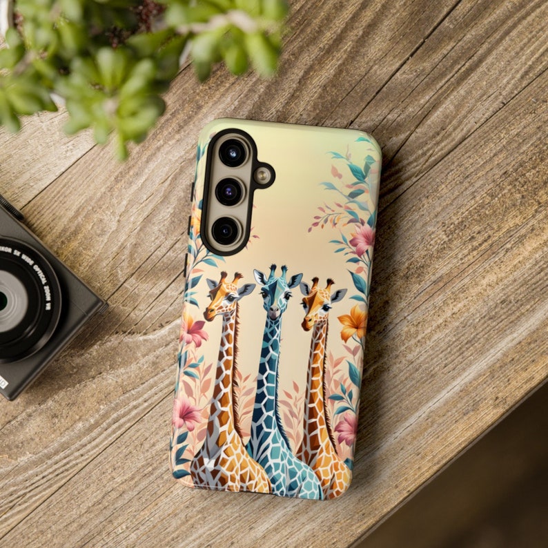 May include: A phone case with a colorful floral design and three giraffes with long necks. The giraffes are in shades of brown, blue, and orange.