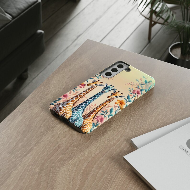 May include: A phone case with a colorful design featuring three giraffes with their heads tilted back, surrounded by flowers and leaves. The case has a camera cutout and a light brown background.