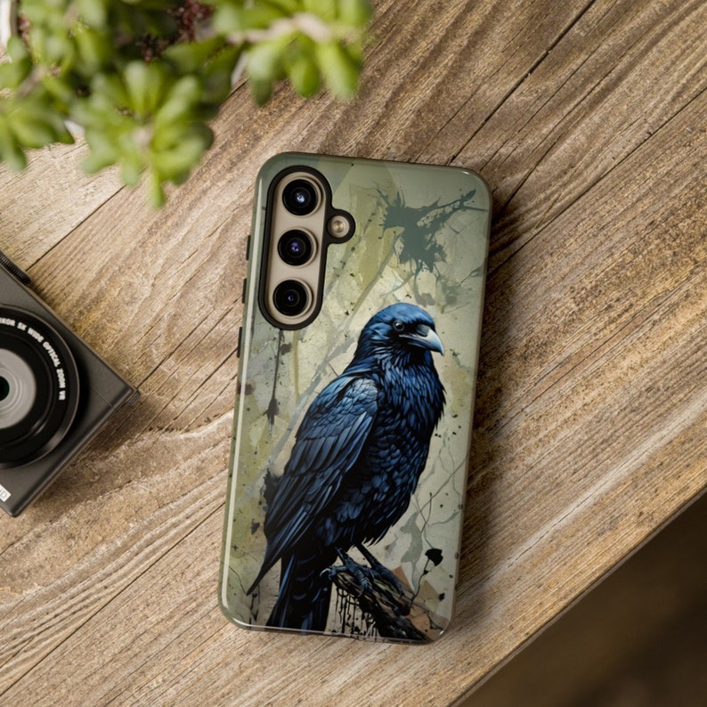 May include: A black phone case with a raven illustration on a green and brown background. The case has a triple camera lens.