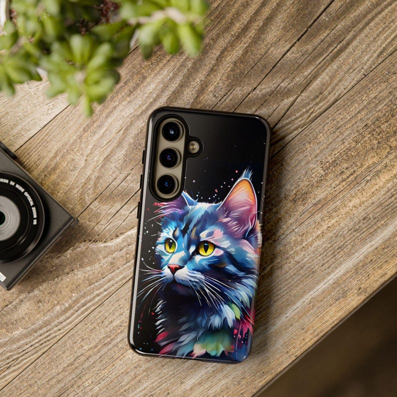May include: A black phone case featuring a colourful illustration of a cat's head. The cat has blue, green and purple fur with yellow eyes. The case has a camera lens cutout with three lenses.
