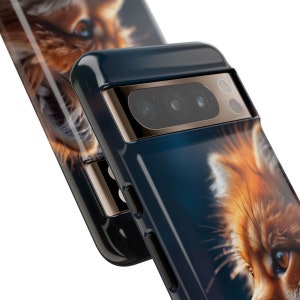 以下が含まれることがあります： 赤いキツネの顔のクローズアップ画像がプリントされた黒い携帯電話ケース。ケースはカメラレンズの周りに金色のトリムがあります。