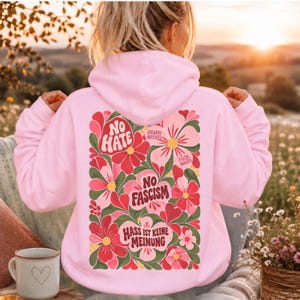 Könnte beinhalten: Ein hellrosa Kapuzenpullover mit einer großen, bunten Blumengrafik auf der Rückseite. Das Design zeigt rote, rosa und grüne Blumen mit dem Text NO HATE, NO FASCISM, GEGEN RECHTS, FUCK NAZIS und HASS IST KEINE MEINUNG auf dem Stoff.