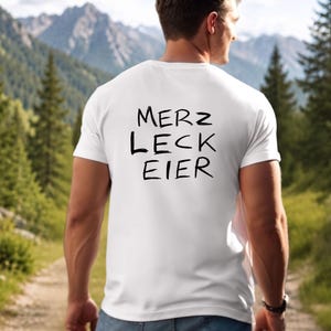 May include: A white short-sleeved t-shirt featuring the black text MERZ LECK EIER printed on the back. The shirt has a crew neckline and a standard fit, constructed from a plain white fabric that displays the bold, dark lettering clearly.