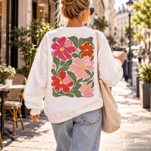 Könnte beinhalten: Ein weißes Langarm-Sweatshirt mit einer großen, bunten Blumengrafik auf der Rückseite. Das Design zeigt rosa, rote und orangefarbene Blumen mit grünen Blättern sowie den Text FUCK AFD, NO HATE, GEGEN RECHTS und FUCK NAZIS.
