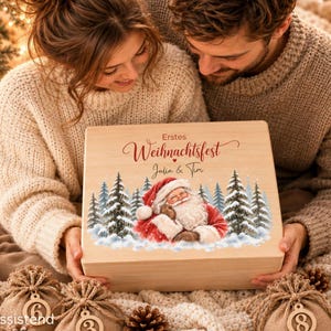 Peut inclure: Une boîte rectangulaire en bois clair avec une illustration imprimée du Père Noël entouré de sapins enneigés. Le texte "Erstes Weihnachtsfest" et "Julia & Tim" est imprimé en écriture rouge et noire sur la surface supérieure.