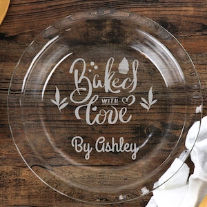 May include: A clear glass pie plate with the words "Baked with Love By Ashley" etched in a decorative font. The plate has a simple, elegant design, perfect for baking and serving homemade pies.