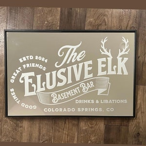 May include: A rectangular sign with a black frame features the text "The Elusive Elk Basement Bar" in white. The sign includes the text "Drinks & Libations" and "Colorado Springs, CO". The sign also includes the text "ESTD 2024" and "Great Friends Good Times".