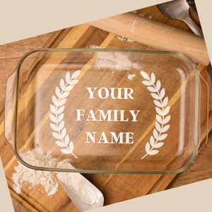 May include: A clear glass baking dish with the text "YOUR FAMILY NAME" inside a white laurel wreath design. The dish sits on a wooden cutting board with flour and a scoop. A rolling pin and spatula are in the background.