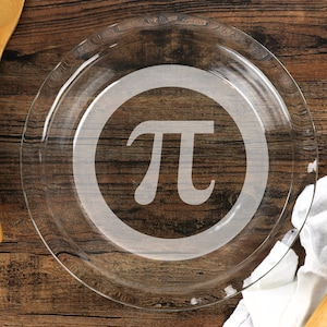 May include: A clear glass pie dish with a circular design and the Greek letter pi etched in the center. The pie plate sits on a wooden surface, showcasing its transparent quality and mathematical theme.