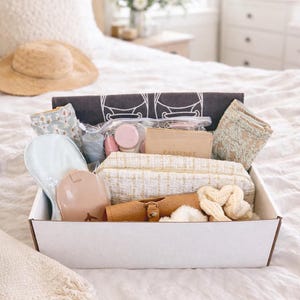 May include: A white box filled with travel accessories, including a straw hat, eye mask, passport holder, cosmetic bag, and scrunchies. The items are in neutral tones of beige, cream, and blush, suggesting a theme of relaxation and travel.