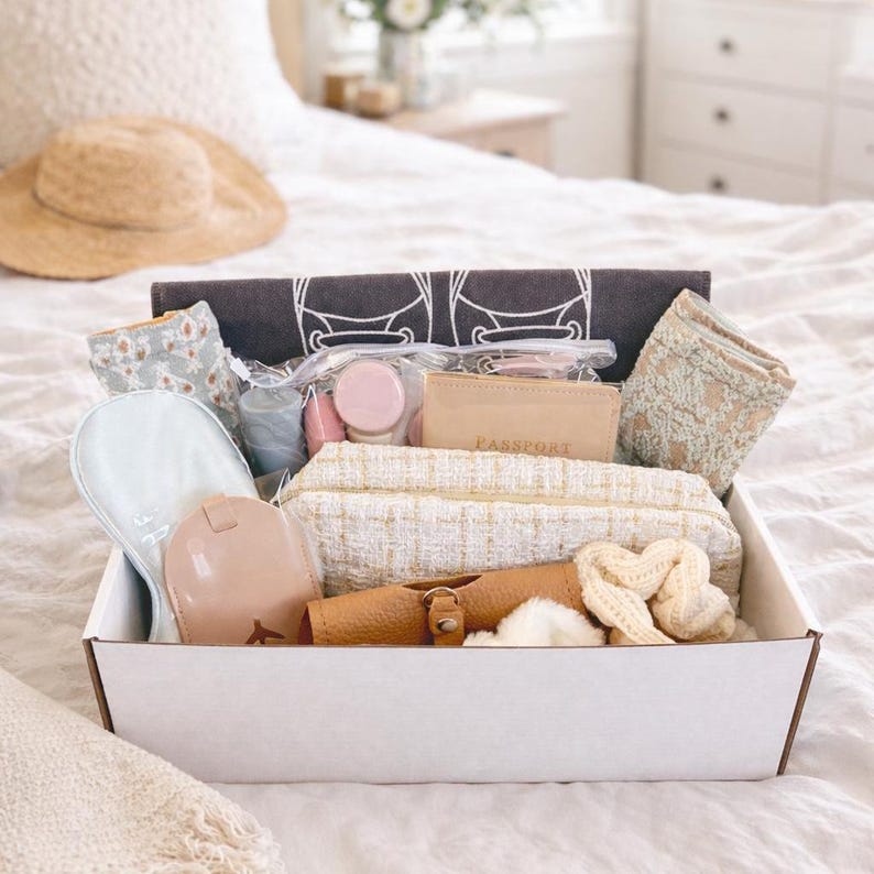 May include: A white box filled with travel accessories, including a straw hat, eye mask, passport holder, cosmetic bag, and scrunchies. The items are in neutral tones of beige, cream, and blush, suggesting a theme of relaxation and travel.