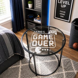May include: A round side table with a black metal frame and a clear glass top. The glass surface features white printed text that reads "JAMES" above "GAME OVER" in a pixelated font, accompanied by a game controller icon and a pac-man character.