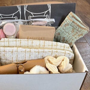 May include: A gift box filled with travel accessories. Includes a passport holder, cosmetic bag, socks, and a small leather item. The cosmetic bag has a white and gold tweed pattern. The passport holder is tan with the word "PASSPORT" embossed on it.