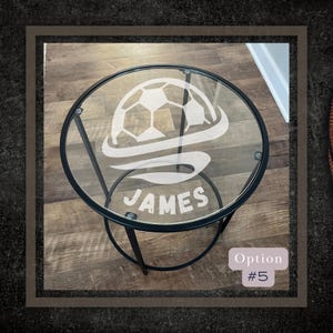 May include: A black metal side table with a round glass top. The glass top has a white soccer ball design with a white swirl and the name "JAMES" etched into the glass.
